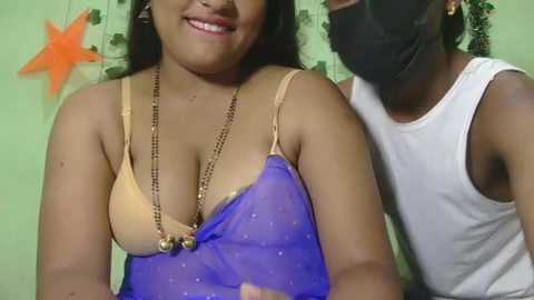 Video of a smiling woman with medium brown skin and medium-length black hair, wearing a sheer blue dress with a beige bra and gold necklace. A man in a white tank top and black face mask stands behind her.