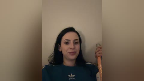 Video of a young woman with long black hair, fair skin, and a slender build, wearing a dark blue Adidas sweatshirt, standing against a plain beige wall, holding a light-colored cane.