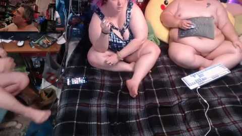 Video of two plus-sized, fair-skinned women with pink hair, one in a blue swimsuit, sitting on a plaid bed playing video games, surrounded by cluttered room.