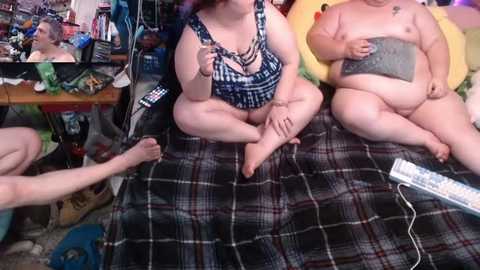 Video of three overweight, fair-skinned individuals in a cluttered room; one wearing a black plaid bikini, two nude. Background features a messy space with a TV, games, and a cat.