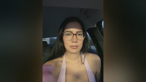 Video of a young woman with long, straight black hair, fair skin, and large glasses, wearing a pink halter top, seated in a dimly lit car.
