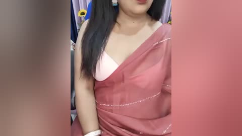 Video of an Asian woman with long black hair, wearing a pink sari with a white bra visible, against a backdrop of floral curtains.