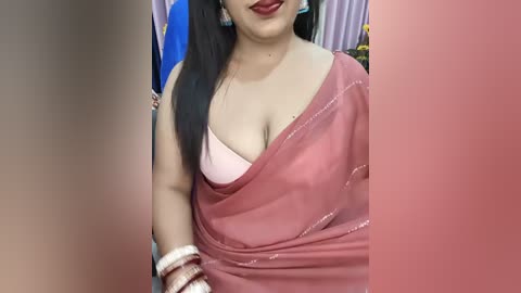 Video of a South Asian woman with medium skin tone, long black hair, and red lipstick, wearing a pink sari with a white bra peeking through, adorned with gold bangles. Background shows a blurred blue flag and yellow flowers.