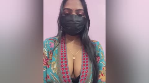 Media: A video of a South Asian woman with medium-dark skin, long black hair, wearing a black face mask, floral-patterned robe, and a low-cut top, against a pink background.