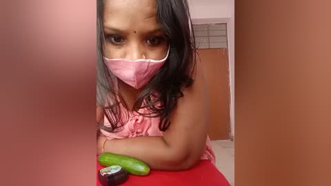 Video of a young Indian girl with medium brown skin and long black hair, wearing a pink face mask and pink top, sitting on a red table, holding a cucumber. Background shows a peach wall and a brown door.