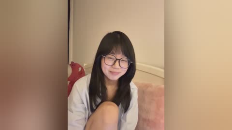 A video of an East Asian woman with straight black hair, wearing glasses, a light-colored shirt, and sitting on a pink couch in a softly lit room with beige walls.