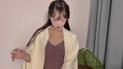 A video of a young East Asian woman with long brown hair, wearing a beige cardigan over a dark brown top, holding a stuffed toy, in a plain indoor setting.