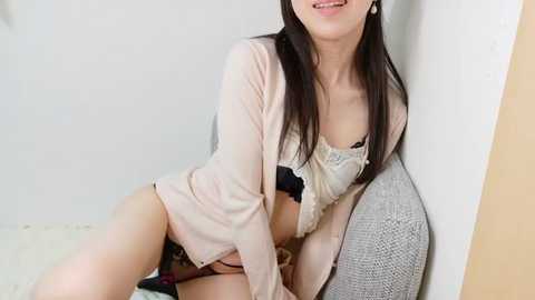 A video of a young East Asian woman with long black hair, wearing a light pink robe and black lingerie, seated against a white wall, smiling.