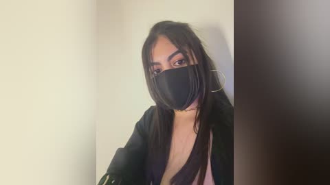 Video of a young woman with medium skin tone and long dark hair, wearing a black face mask, black jacket, and a white top, against a plain wall background.