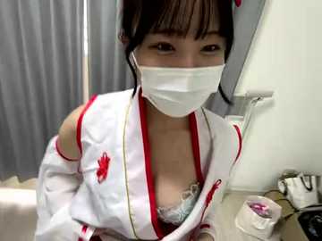 A video of an East Asian woman in a revealing white nurse outfit with red accents, showing cleavage, and a surgical mask. She is holding a mirror in a dimly lit room.
