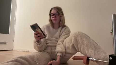 Video of a blonde woman with glasses, wearing a cream-colored, fluffy bathrobe, sitting on a wooden floor, reading a smartphone.