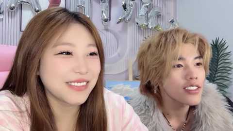 Video of two young Asian women with long hair, one in pink, the other in white fur, smiling, sitting in a modern bathroom with silver fixtures on a white wall.
