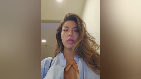 Video of a young woman with light brown skin, long wavy brown hair, wearing a light blue shirt over a tan top, looking serious, standing indoors with beige walls and recessed lighting.
