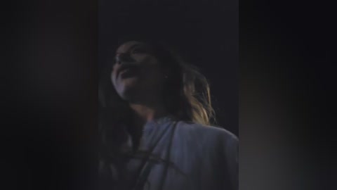 A dimly lit video of a woman with long brown hair, wearing a white shirt, looking upward in a contemplative or emotional state, with a dark, shadowy background.