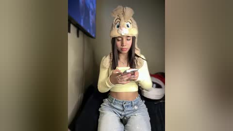 Video of a young woman with light brown skin, long dark hair, wearing a yellow crop top, light blue jeans, and a fuzzy yellow beanie with cartoon eyes, playing a smartphone indoors.