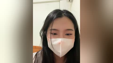 Video of an Asian woman with long black hair and fair skin, wearing a white face mask indoors. She has a neutral expression, and the background shows a white wall and a wooden chair.