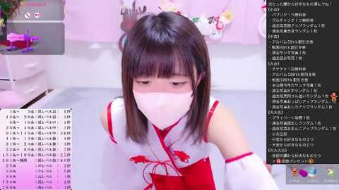 A video of an Asian woman with straight black hair and a white mask, wearing a red and white traditional Japanese kimono, kneeling on a white floor in a room with pink decor and floral arrangements.
