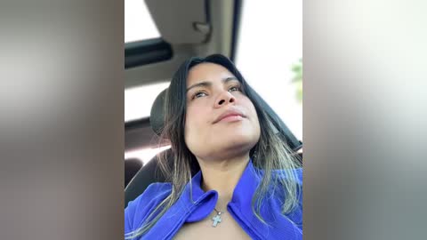 A video of a Latina woman with long, dark hair and light brown skin, wearing a blue shirt, sitting in a car. She has a neutral expression and a small necklace. The car's interior is visible in the background.