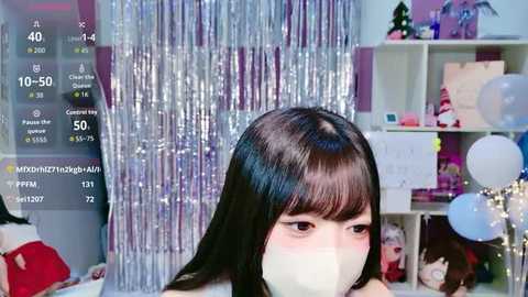 Video of a young East Asian woman with long black hair, wearing a white mask, standing in a festive room with silver tinsel, white balloons, and colorful decorations.