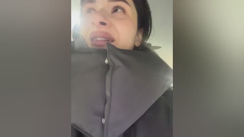Video of a woman with fair skin and dark hair, wearing a grey, puffy jacket, looking upwards with a surprised expression, possibly in a dimly lit room.