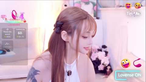 Video of a young Asian woman with long brown hair in a ponytail, wearing a white tank top, sitting in a playful bedroom setting.