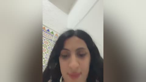 Media: Video of a young woman with long, dark hair, fair skin, and light makeup, smiling, in a room with a tiled wall and white ceiling.