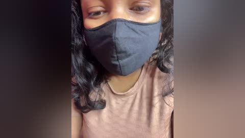 Video of a young girl with medium brown skin and long, wavy black hair. She wears a black face mask, a pink t-shirt, and a dark background.