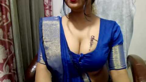 Video of a woman with medium skin tone, wearing a blue sari with gold trim, sitting indoors. She has medium-sized breasts and a tattoo on her chest.