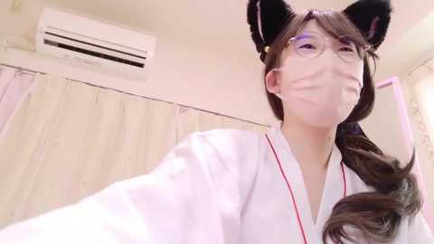 Video of a smiling Asian woman wearing cat ears, a white robe, and glasses, in a hospital room with a white curtain, air conditioner, and pink floral wallpaper.