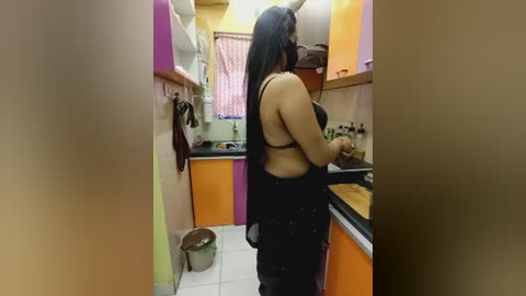Video of a woman with long black hair, wearing a black halter top and black pants, cooking in a small, brightly colored kitchen with orange and purple cabinets.