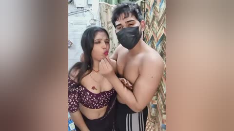 Video of a topless man with a mask and a woman in a patterned crop top and mask, kissing in a backyard with a bamboo fence and concrete floor.