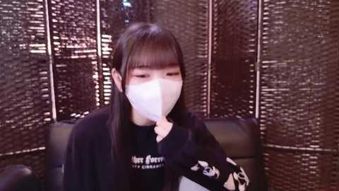 Video of an East Asian woman with long black hair, wearing a black long-sleeve shirt, white face mask, and seated indoors, surrounded by dark, woven bamboo screens.