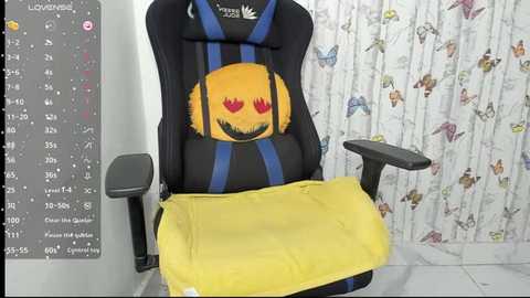 Video of a gamer chair with a plush, yellow smiling face pillow, blue and black striped cushion, and a yellow blanket draped over the armrest. Background features a colorful butterfly-patterned wallpaper and a calendar.