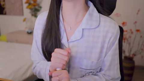 Video of a young woman with long black hair wearing a light blue, open-collared shirt, seated indoors with blurred background featuring a white bed, vase of flowers, and decorative items.