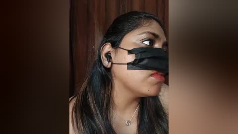 Media: Video of a young woman with medium-brown skin, long black hair, wearing a black face mask and a light-colored top, looking pensive against a wooden backdrop.