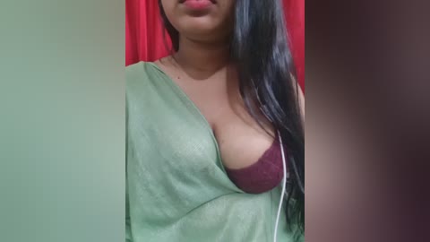 Video of a woman with medium skin tone and long black hair, wearing a green cloth draped over her left shoulder, revealing a burgundy bra. Background features red curtains and blurred walls.
