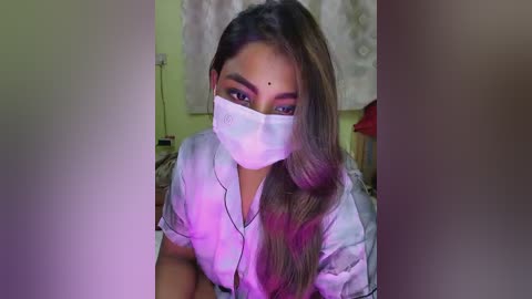 Video of a woman with long, dark hair, wearing a white mask, purple shirt, and headband, indoors with a white wall and blurred background.
