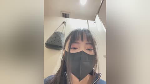Video of an Asian woman with long black hair, wearing a black face mask, blue jacket, and grey purse, taken indoors with beige walls and a ventilation grille visible in the background.