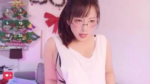 Video of a young Asian woman with fair skin, glasses, and ponytail, wearing a white sleeveless top, smiling in a cozy, decorated room with a Christmas tree.