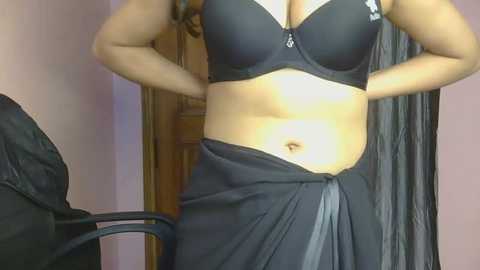 Video of a woman with medium brown skin, wearing a black bra and a black sarong, standing indoors near a wooden door.