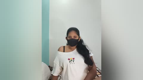 A video of a young woman with long black hair in a high ponytail, wearing a black face mask, white T-shirt with colorful floral print, and sitting in a room with white walls and a checkered floor.