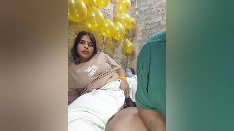 Media: A video shows a woman with dark skin and long hair, wearing a beige sweatshirt and white pants, sitting among yellow balloons against a brick wall. Another person is partially visible in the foreground.