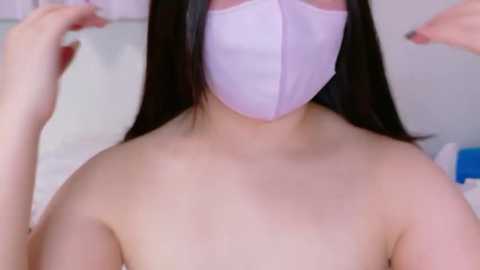 Video of a woman with long black hair, wearing a pink face mask, topless, with her hands raised in front of her.