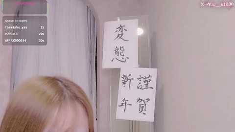 Video of a blonde woman in a room with Japanese calligraphy on the wall; her hair is slightly out of focus.