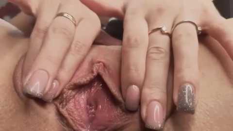 Media: A close-up video of a fair-skinned woman's vulva, with her fingers spreading the labia to reveal the inner pink tissue, adorned with glittery nail polish and a silver ring.