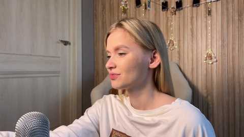 Video of a blonde woman with fair skin wearing a white T-shirt, sitting in front of a microphone, in a cozy, wooden-paneled room with fairy lights.