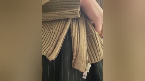 Video of a person's torso, partially dressed in a beige knit sweater and black pinstriped skirt. The sweater's ribbed texture contrasts with the smooth skirt fabric. The background is a plain beige wall.