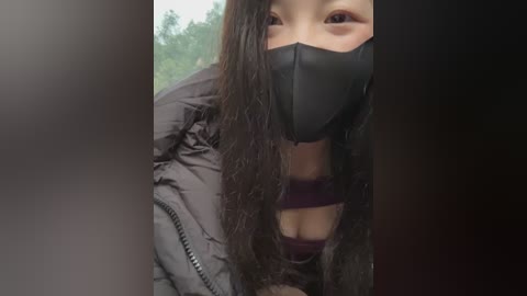 Video of an East Asian woman with long, wet black hair, wearing a black face mask, puffer jacket, and a dark bra, outdoors with blurred greenery in the background.