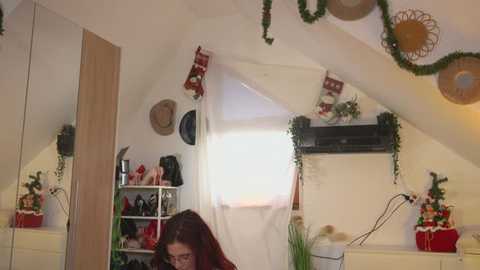 Video of a cozy attic bedroom with Christmas decorations, including a festive tree and wreath, a woman in a red sweater, and a white bed covered in a white sheet.