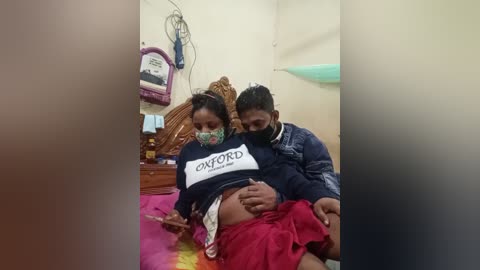 A video captures a pregnant woman in a small, modest room. She is wearing a black sweatshirt with the word \"OXFORD\" and has a mask on. The room is cluttered with various items, including a bed and a wall clock.